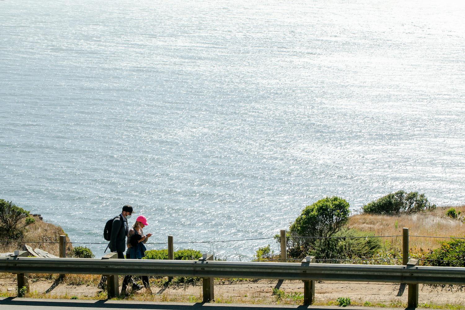 Man and woman walking along a dirt trail next to a roadside with the Pacific Ocean in the background.